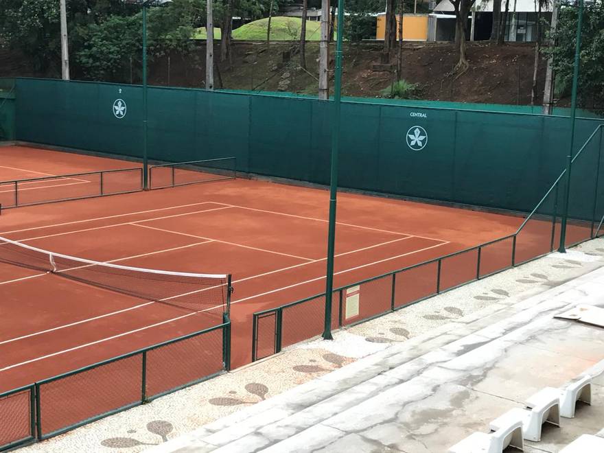 Quadra central de saibro com tela de fechamento verde e o brasão do clube aplicado.